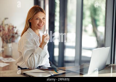 Smiling business woman working on laptop, making notes in cozy coworkign space interior Stock Photo