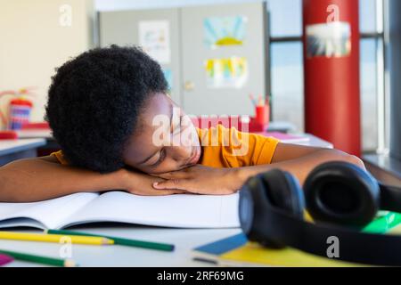Tired biracial schoolboy sitting at desk and lying on books in classroom Stock Photo