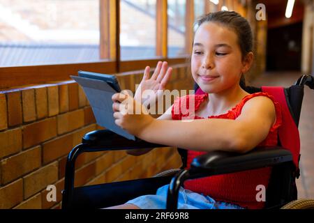 Happy caucasian schoolgirl sitting in wheelchair and using tablet at school corridor Stock Photo