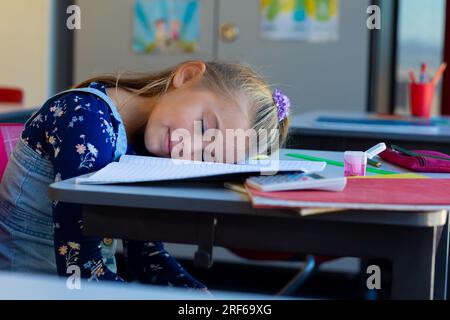 Tired caucasian schoolgirl sitting at desk and lying on books in classroom at elementary school Stock Photo
