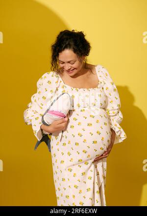 Cheerful curly multi-ethnic pregnant woman smiling, holding a plush shark toy, isolated over yellow studio background. Maternity and pregnancy concept Stock Photo