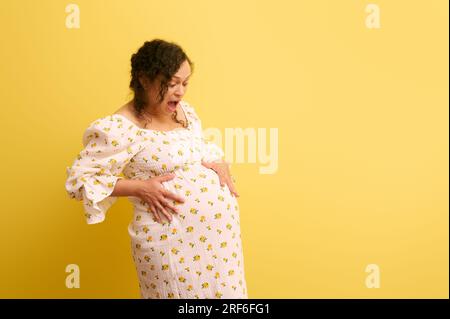 Curly ethnic pregnant woman, gravid female, mother to-be holding belly, crying and shouting, isolated on yellow background. Easy delivery, pregnancy a Stock Photo