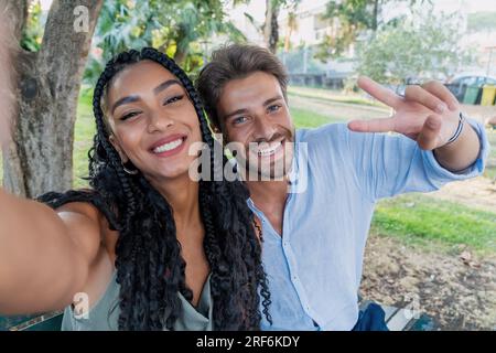 Young African American woman with braided hair taking a selfie with her best Caucasian friend. They are sitting on a park bench, enjoying their time t Stock Photo