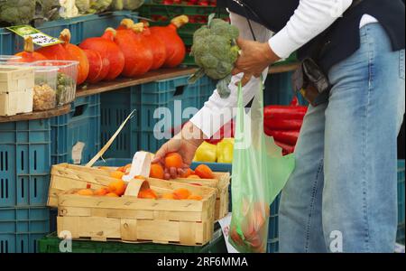 A woman, visitor at the Naplavka farmers market buying fresh fruit and vegetables, broccoli and picking apricots from a wooden basket. Stock Photo