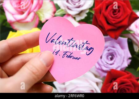 FOTOMONTAGE, Hand hält herzförmigen Zettel mit Aufschrift Valentinstag 14. Februar vor einem Strauß Rosen