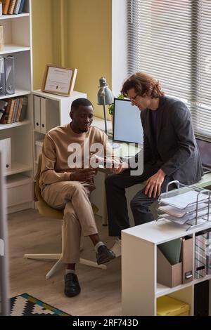 Vertical full length portrait of black young man talking to colleague while working in office by window Stock Photo