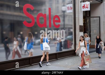 People out shopping walk past a large scale sale sign in red and silver for major high street clothing brand H&M outside their flagship store on the corner of Oxford Street and Regent Street on 9th July 2023 in London, United Kingdom. Oxford Street is a major retail centre in the West End of the capital and is Europes busiest shopping street with around half a million daily visitors to its approximately 300 shops, the majority of which are fashion and high street clothing stores. Stock Photo