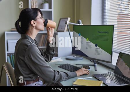 Side view portrait of young woman drinking coffee in office while working on development strategies by deadline Stock Photo