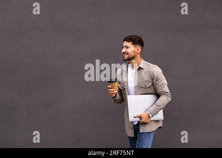 Cheerful middle eastern young businessman in glasses with laptop and coffee to go walking by street towards modern office building, going to job in th Stock Photo