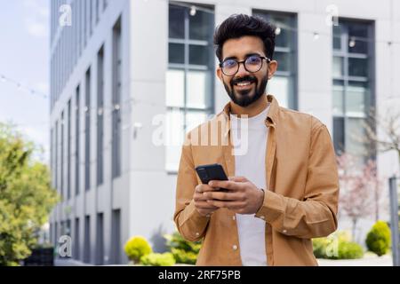 Successful young Hindu man walking outside office building, engineer software developer programmer smiling and happy using test app on phone, happy satisfied with result. Stock Photo