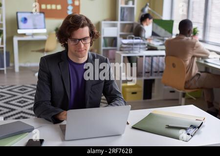 Portrait of young long haired businessman wearing glasses using laptop at office workplace and smiling, copy space Stock Photo