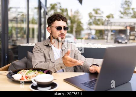 Handsome smiling modern man waving hand to laptop web camera while chatting with family online at street cafe. Video call concept Stock Photo