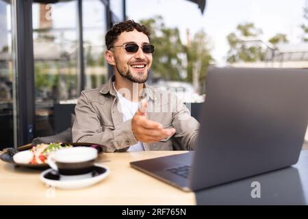 Handsome smiling modern man waving hand to laptop web camera while chatting with family online at street cafe. Video call concept Stock Photo