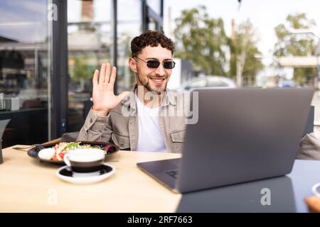 Handsome smiling modern man waving hand to laptop web camera while chatting with family online at street cafe. Video call concept Stock Photo