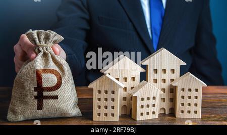 A businessman puts a russian ruble money bag near the houses. Investments in real estate assets. Municipal budget of the city. Official. Construction Stock Photo