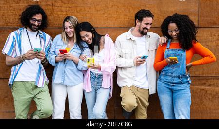 Diverse young confident people smiling and using their phones outside in a city while leaning against a wooden wall. Multiracial group of friends Stock Photo