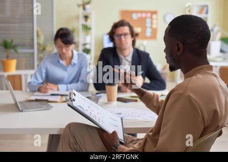 Side view portrait of black young man holding clipboard while leading business meeting with team in office and discussing project strategy, copy space Stock Photo