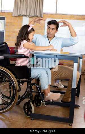 Happy biracial male teacher helping schoolgirl sitting in wheelchair in class at elementary school Stock Photo