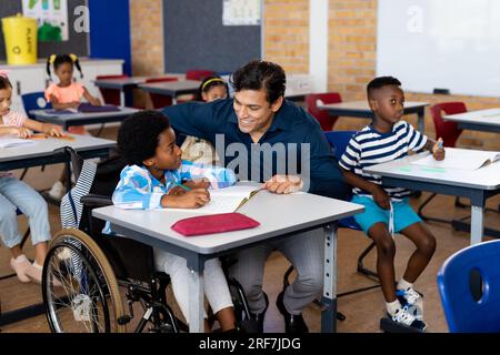 Happy biracial male teacher with boy in wheelchair in class at elementary school Stock Photo