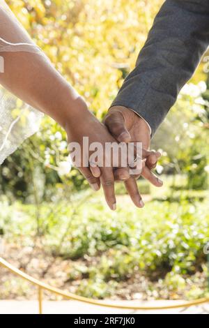 Midsection of senior biracial bride and groom holding hands at sunny outdoor wedding ceremony Stock Photo