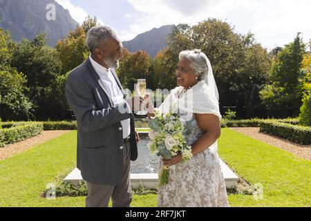 Happy senior biracial bride and groom drinking champagne toast in garden at sunny wedding ceremony Stock Photo