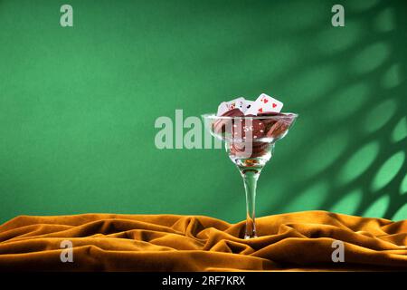 Red plastic poker chips lie in a glass margarita glass on a brown velvet cloth.  A fan of small playing cards on the rim of a glass, like a cocktail d Stock Photo