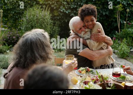 Affectionate senior man embracing his African American grandson while sitting by dinner table served with healthy and tasty homemade food Stock Photo