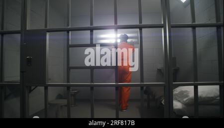 African American prisoner in orange uniform looks at barred window and leans on prison cell bars. Depressed inmates in detention center. Murderers serve imprisonment term in jail cell. Stock Photo