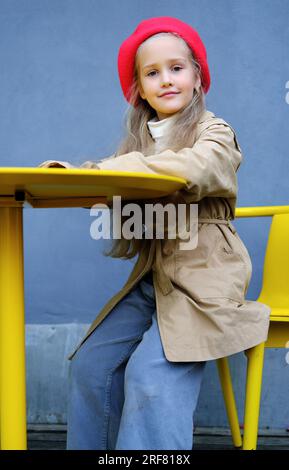 Beautiful little girl in a trench coat, red beret and jeans posing while sitting at the table of a street cafe. Vertical photo Stock Photo