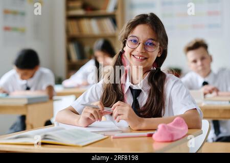 Portrait of schoolgirl in eyeglasses smiling at camera while sitting at her desk at lesson Stock Photo