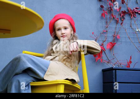 Beautiful little girl in a trench coat, red beret and jeans posing while sitting at the table of a street cafe. Horizontal photo Stock Photo