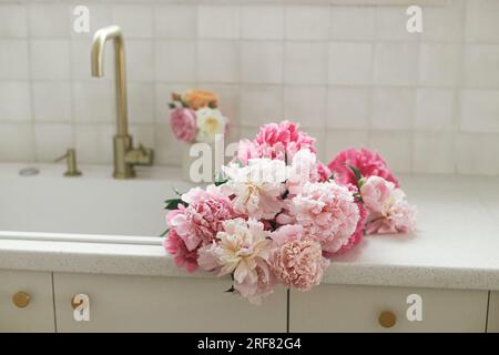 Beautiful peonies in sink on background of brass faucet and white counter in new scandinavian house. Pink peony and roses flowers in modern kitchen in Stock Photo