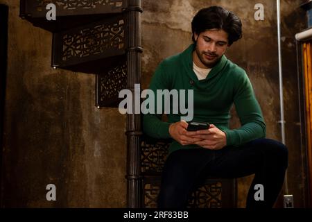 Indian man sitting on stairs and using smartphone at home, copy space Stock Photo