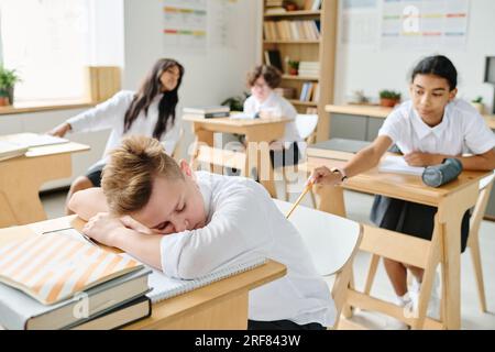 Schoolboy sleeping at desk at lesson while his classmate waking him up Stock Photo