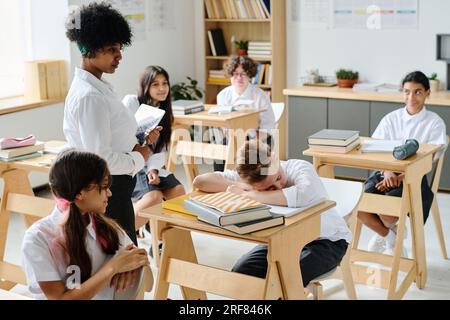 Schoolboy sleeping at his desk during lesson at school while teacher waking him up Stock Photo