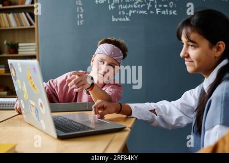 Schoolgirl using laptop while sitting at table and talking to her classmate during lesson Stock Photo