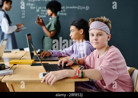 Portrait of schoolboy looking at camera while sitting at desk and studying on laptop with his classmates in background Stock Photo