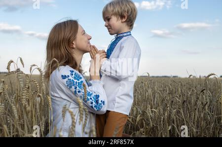 cute boy and a woman in embroidered national clothes are sitting on a field among wheat spikelets. Family, refugees, unity, support, tenderness of mot Stock Photo