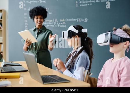 Students using VR glasses while sitting at table and listening to teacher at IT lesson Stock Photo