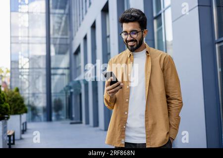 Young successful businessman walking in the city outside office building, hispanic smiling happy with achievement results, holding phone, using online application outdoors. Stock Photo