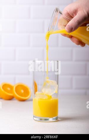A man pours orange juice from a bottle into a tall glass with ice. The concept of freshness and usefulness. In the background is an orange cut in half Stock Photo