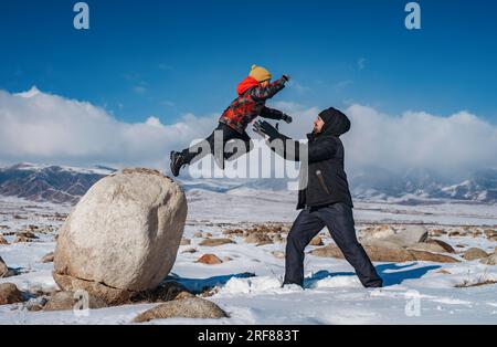 Child jumping on daddy from big wall on mountains background at winter season Stock Photo