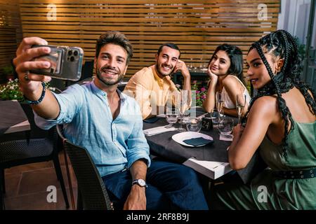 A diverse group of young adults, two Caucasian men, a Caucasian woman, and an African American woman, smiling and taking a selfie on a restaurant terr Stock Photo
