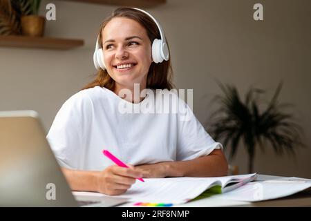 Happy female student in headphones sitting at desk at home doing homework in workbook. Stock Photo