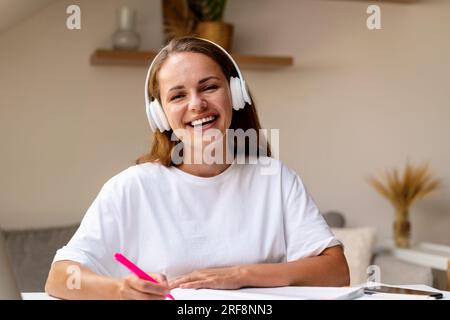 Front view portrait of young laughing female student in headphones wearing white tee shirt doing homework. Study, learning, education. Stock Photo