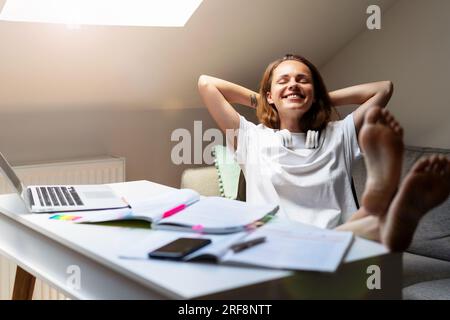 Young woman student resting with hands behind her head and feet on the table after homework done, enjoying and dreaming. Stock Photo