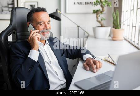 Smiling confident caucasian senior businessman in suit work on laptop, calling by smartphone at table in office Stock Photo