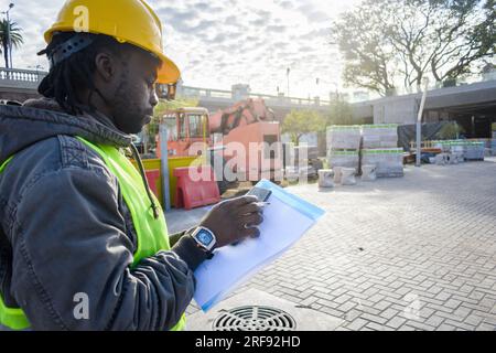 rear view of young black man in yellow safety helmet, vest and beard, construction site supervisor standing using phone, copy space with construction Stock Photo