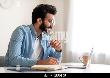 Man looking at laptop screen and writing on paper at dining table in ...