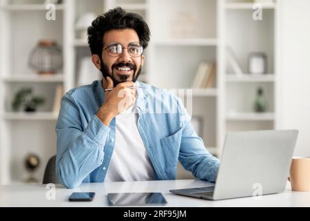 Dreamy Young Indian Man Sitting At Desk With Laptop In Home Office Stock Photo